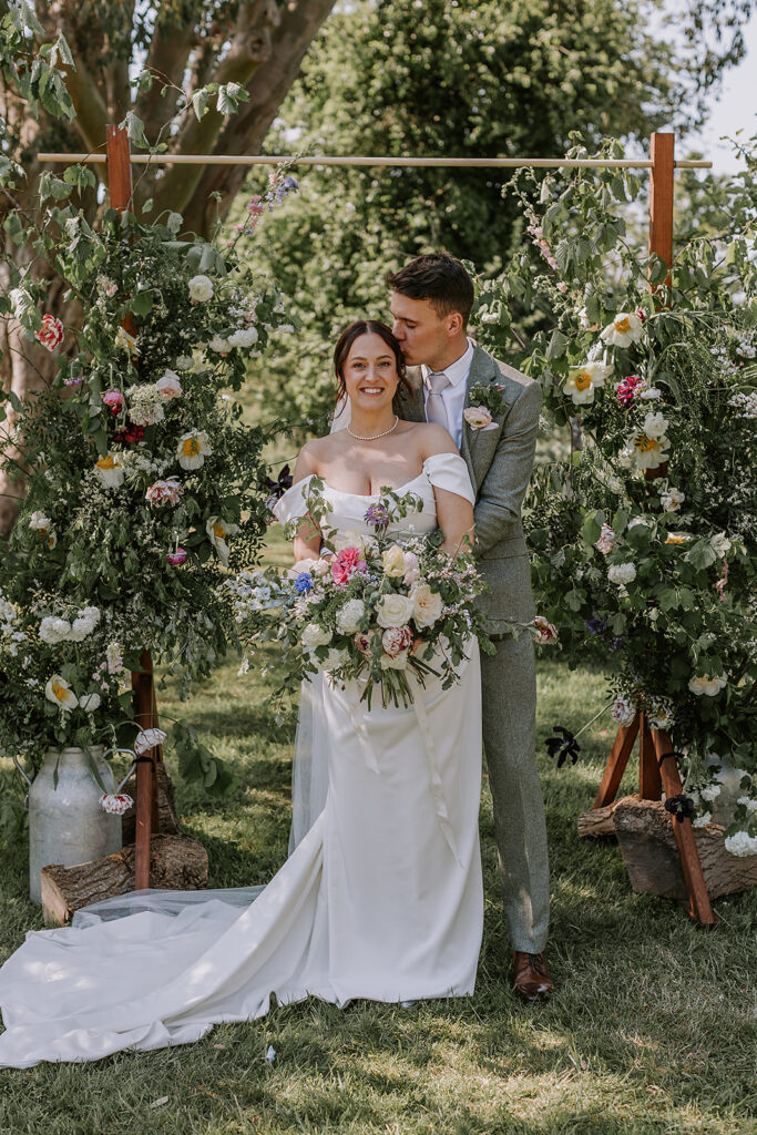 Just married couple under a floral wedding arch in the orchard at The Secret Barn.